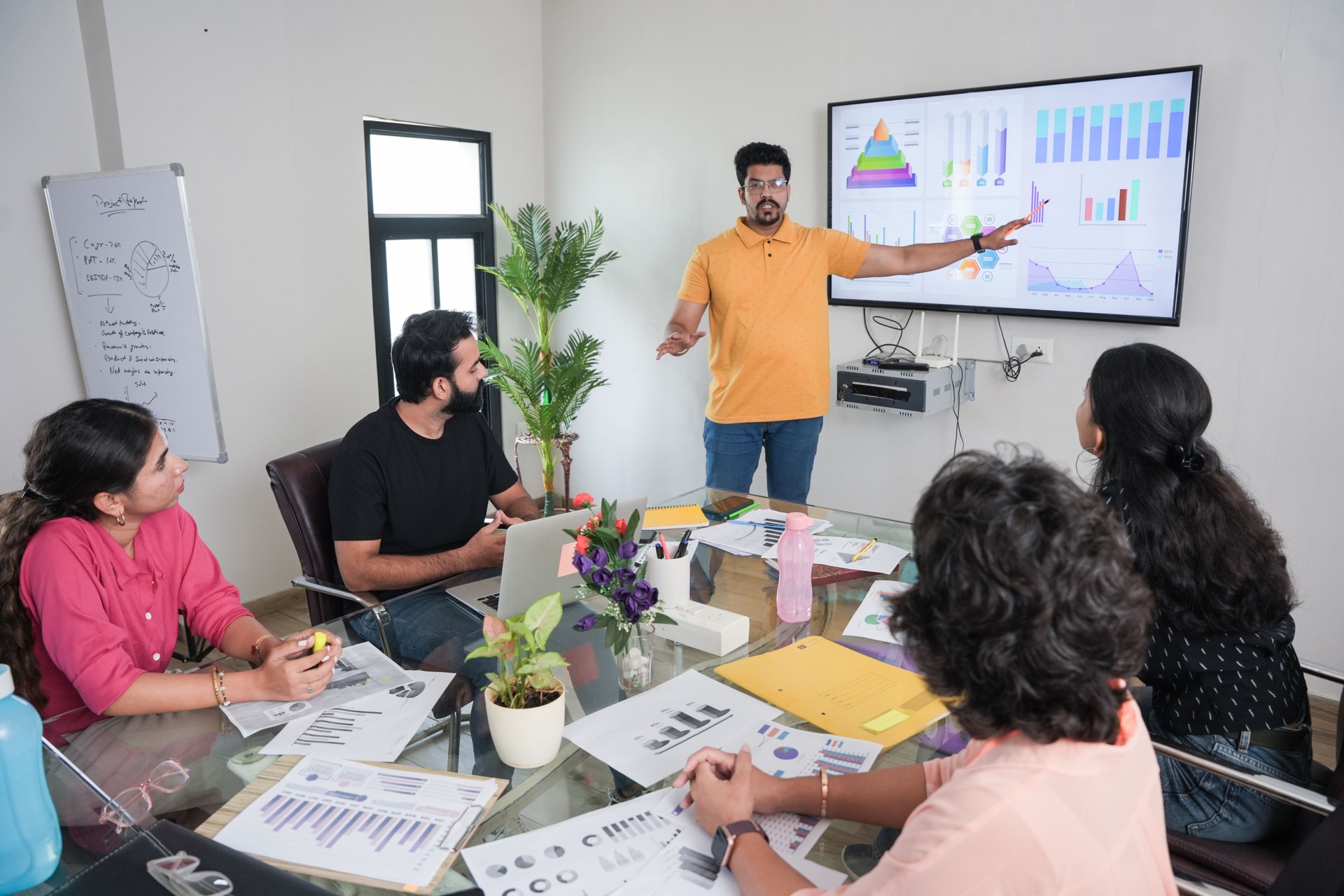 young indian man entrepreneur presents financial data with charts and graphs on wall tv in the business meeting room to colleagues for a start-up project. sales, analysis and strategy concepts. Teamwork